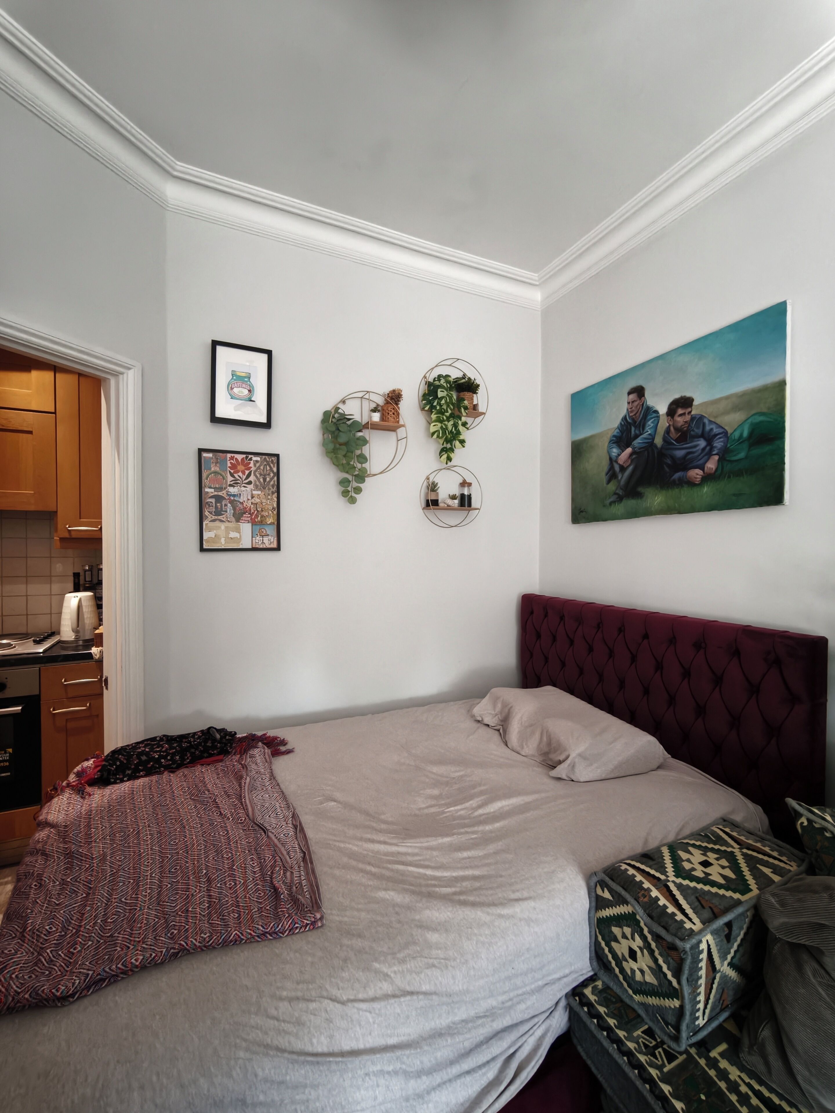 A bedroom with a bed, a headboard, a wall-mounted shelf with plants, and a framed picture.