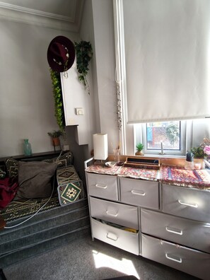 A room with a desk, drawers, a window with blinds, a plant, and a decorative wall hanging. - Cute studio apartment on café street close to music venues and camden (London)