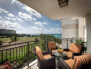 A balcony with wicker furniture, a glass table, and a view of the ocean and a golf course. - Beachfront 2 Condos at Ko Olina Beach Villas · 6 BR · Sleeps 16 (Kapolei)