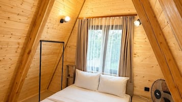 Ein Schlafzimmer mit Holzbalkendecke, einem Bett mit weißer Bettwäsche, einem Fenster mit Vorhängen und einem Standventilator.