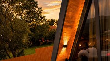 Un bain à remous avec un flotteur en forme de flamant rose, entouré d’une terrasse en bois et d’une structure en verre offrant une vue sur les arbres et un coucher de soleil.
