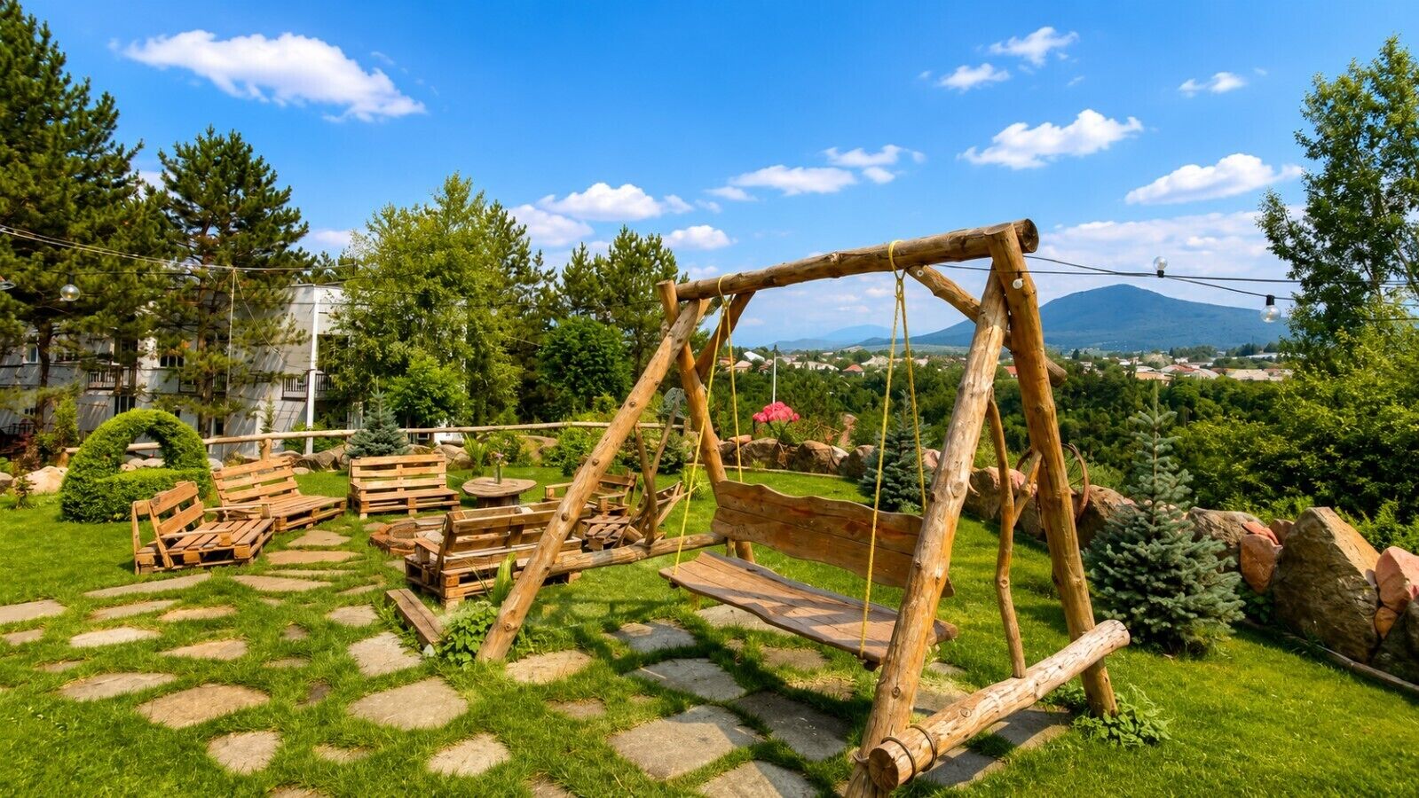 Ein Sitzbereich im Freien mit Holzbänken und -tischen, umgeben von einem gepflegten Rasen und Blick auf ferne Berge.