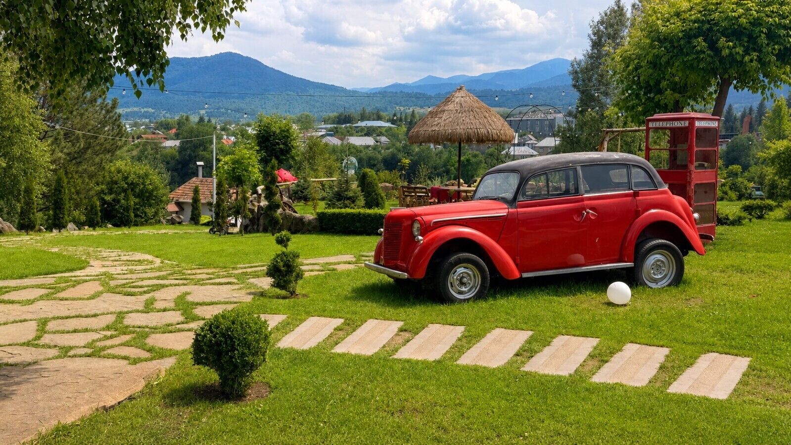 Ein rotes Oldtimer-Auto auf einem gepflasterten Weg in einem üppigen Garten mit Bergblick.