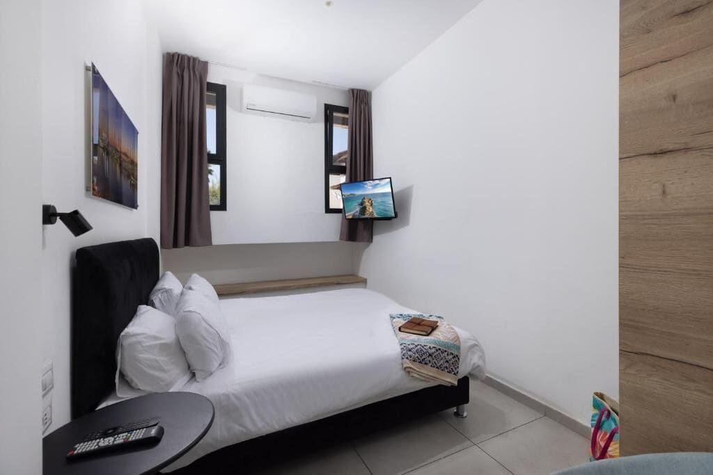 A hotel room with a bed, a flat-screen TV mounted on the wall, a bedside table, and a window with curtains.
