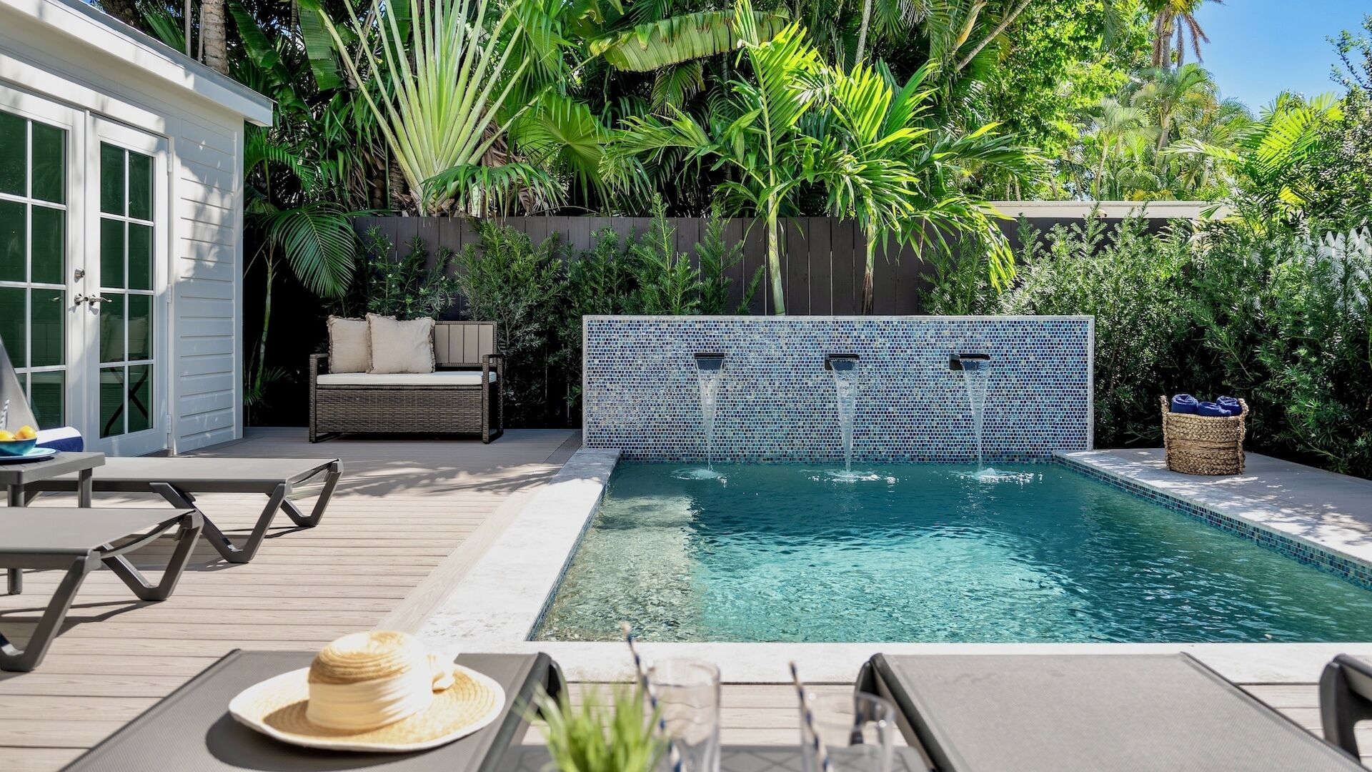 A swimming pool with a tiled wall, surrounded by lush greenery and a wooden deck with outdoor furniture.