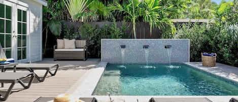 A swimming pool with a tiled wall, surrounded by lush greenery and a wooden deck with outdoor furniture.