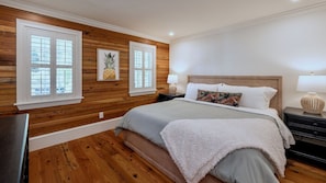 Ein Schlafzimmer mit Holzwandverkleidung, einem Bett mit grauer Bettdecke, zwei Nachttischlampen und einem Bilderrahmen mit einer Ananaszeichnung an der Wand.