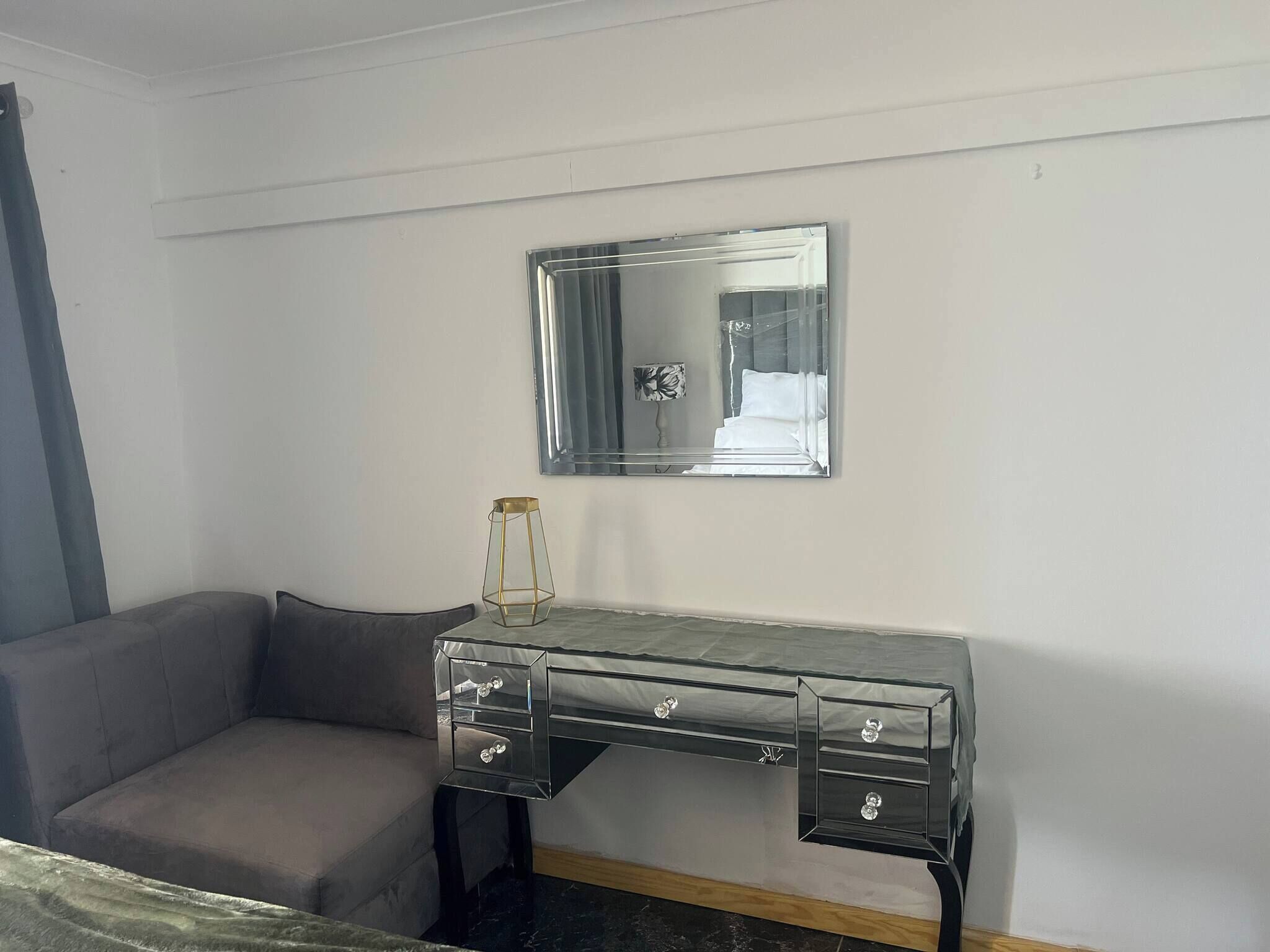 A room with a grey sofa, a mirrored dresser with drawers, and a small table with a lamp.