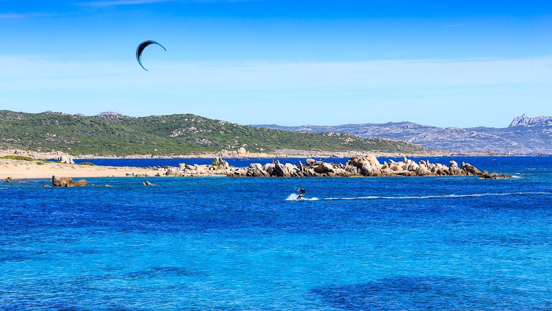 Eine Küstenlandschaft mit klarem, blauem Wasser, einer Person beim Kitesurfen und Felsen.