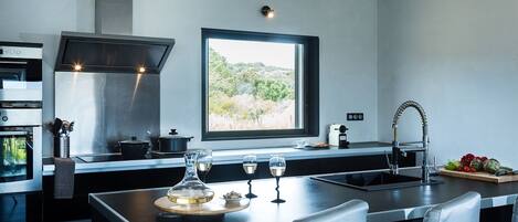 A modern kitchen with a large window, stainless steel appliances, and a central island with glasses and a decorative bowl.