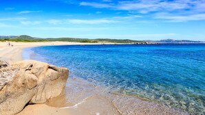 Uma praia com águas azul-claras, uma rocha grande em primeiro plano e uma linha de costa distante com colinas.