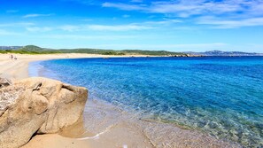 Una playa con agua azul cristalina, una roca grande al frente y una costa distante con colinas.