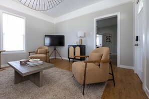 Uma sala de estar com televisão de tela plana, dois poltronas, uma mesa de centro e um abajur.