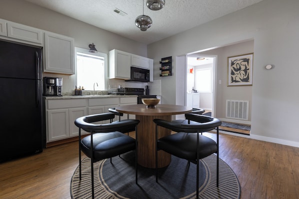 A modern kitchen with white cabinets, a black refrigerator, a microwave, and a coffee maker. There is a round wooden table with four black chairs and a decorative bowl.