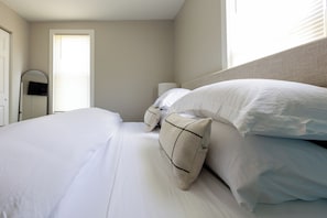 A neatly made bed with white linens and multiple pillows, a bedside table with a lamp, and a window with blinds.