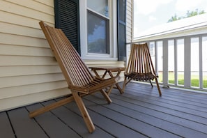 Two wooden Adirondack chairs on a wooden deck with a white railing, a window with black shutters, and a white exterior wall.