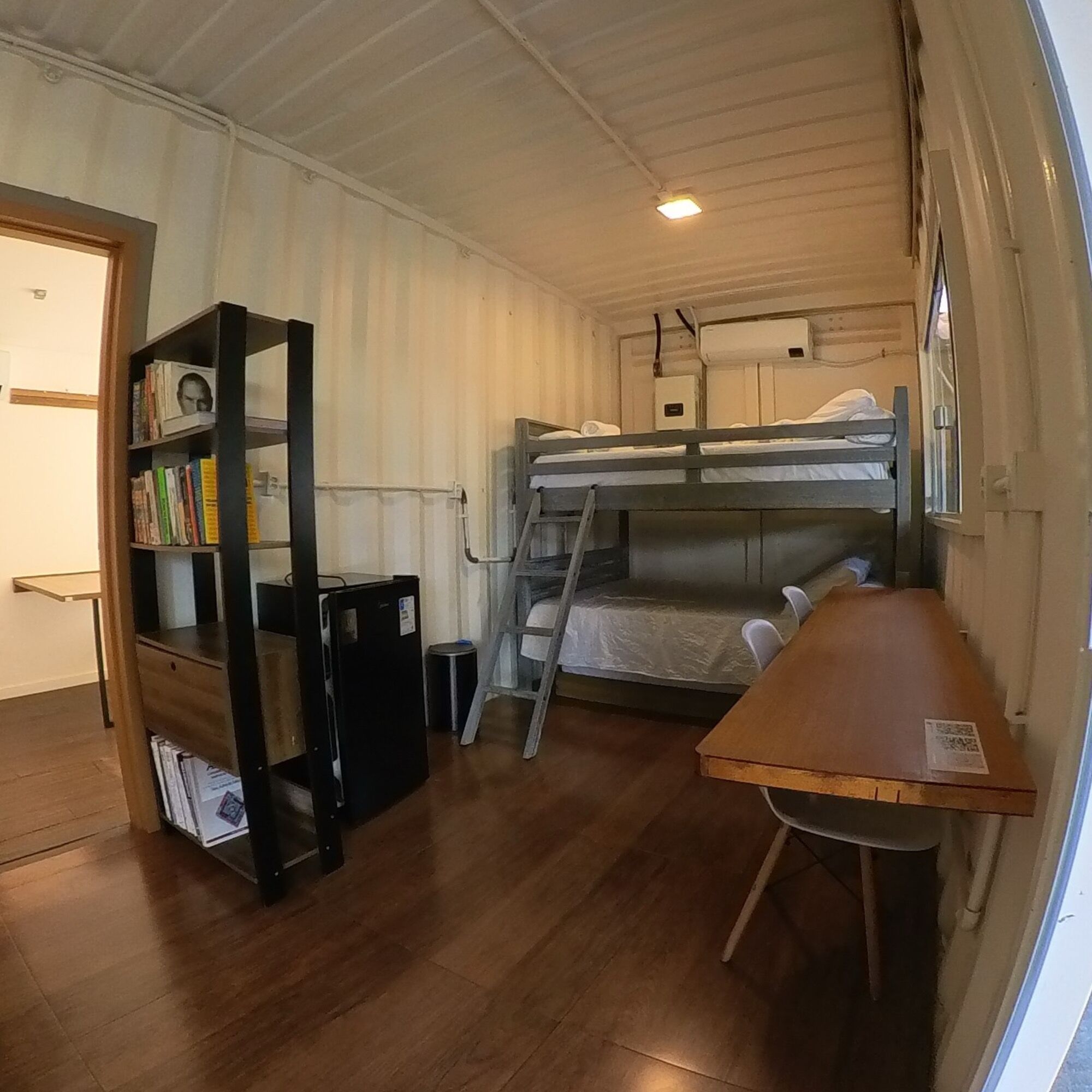 Quarto pequeno com beliche, escrivaninha, estante e escada.