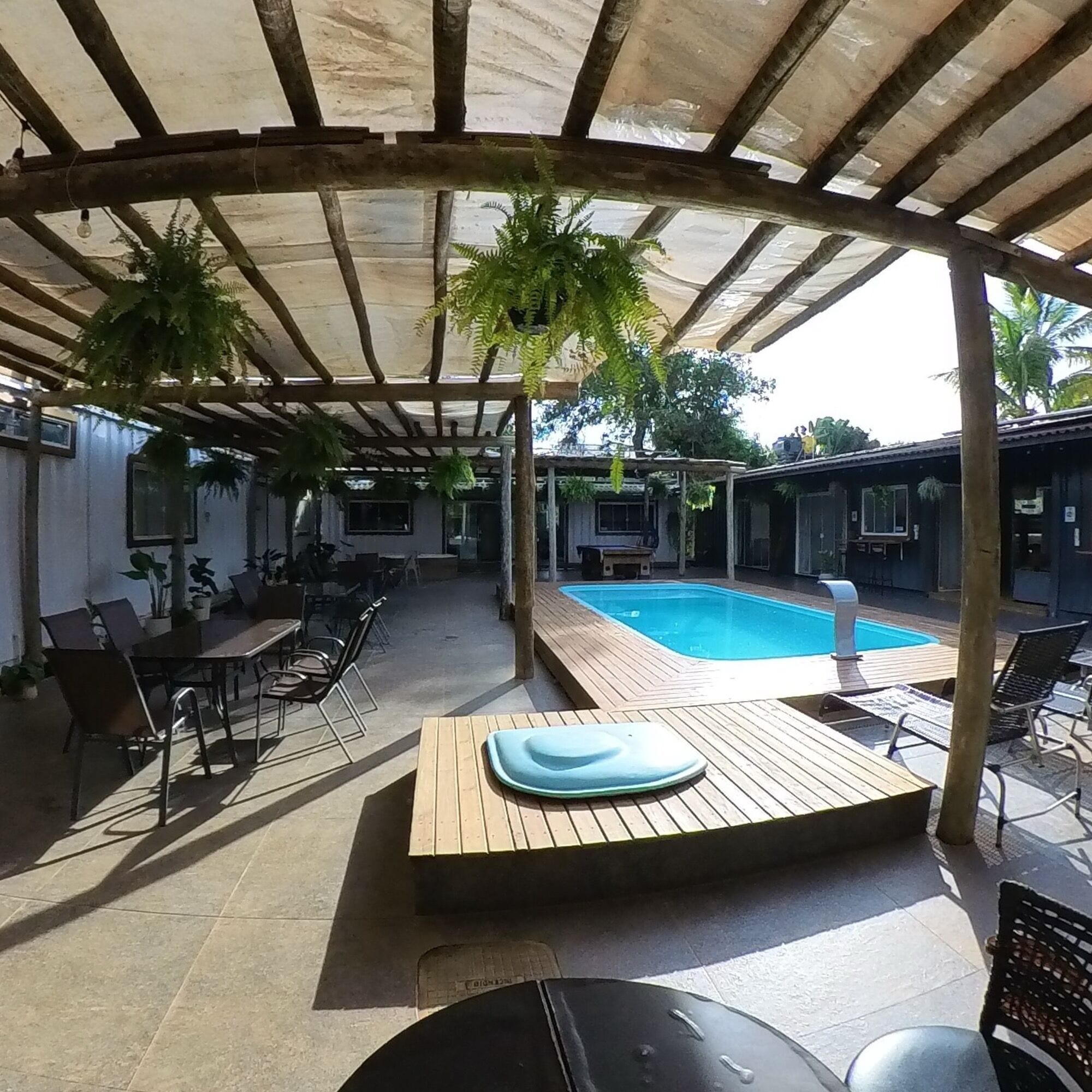 Uma área externa coberta com piscina, móveis de madeira e plantas em vasos.