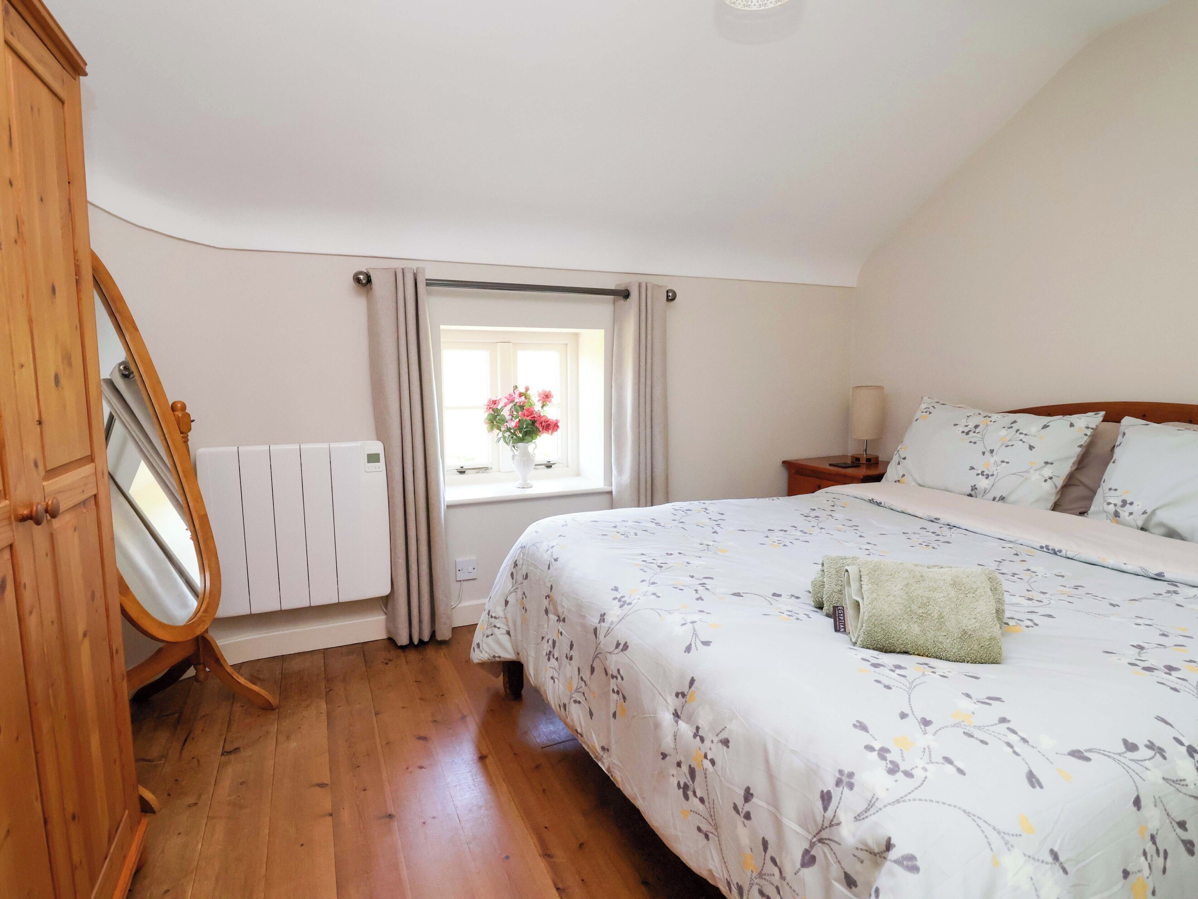 A bedroom with a wooden floor, a bed with floral bedding, a bedside table with a lamp, a window with curtains, and a radiator.