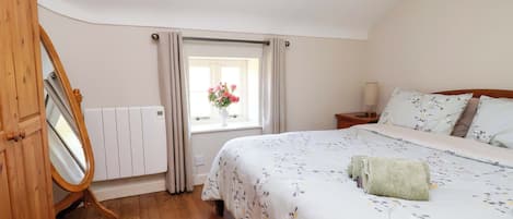 A bedroom with a wooden floor, a bed with floral bedding, a bedside table with a lamp, a window with curtains, and a radiator.