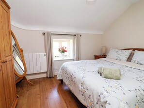 A bedroom with a wooden floor, a bed with floral bedding, a bedside table with a lamp, a window with curtains, and a radiator. - Pen Y Grisiau (Mold)