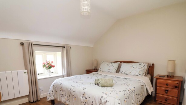 A bedroom with a wooden floor, a bed with floral bedding, a bedside table with a lamp, and a window with curtains. - Pen Y Grisiau (Mold)