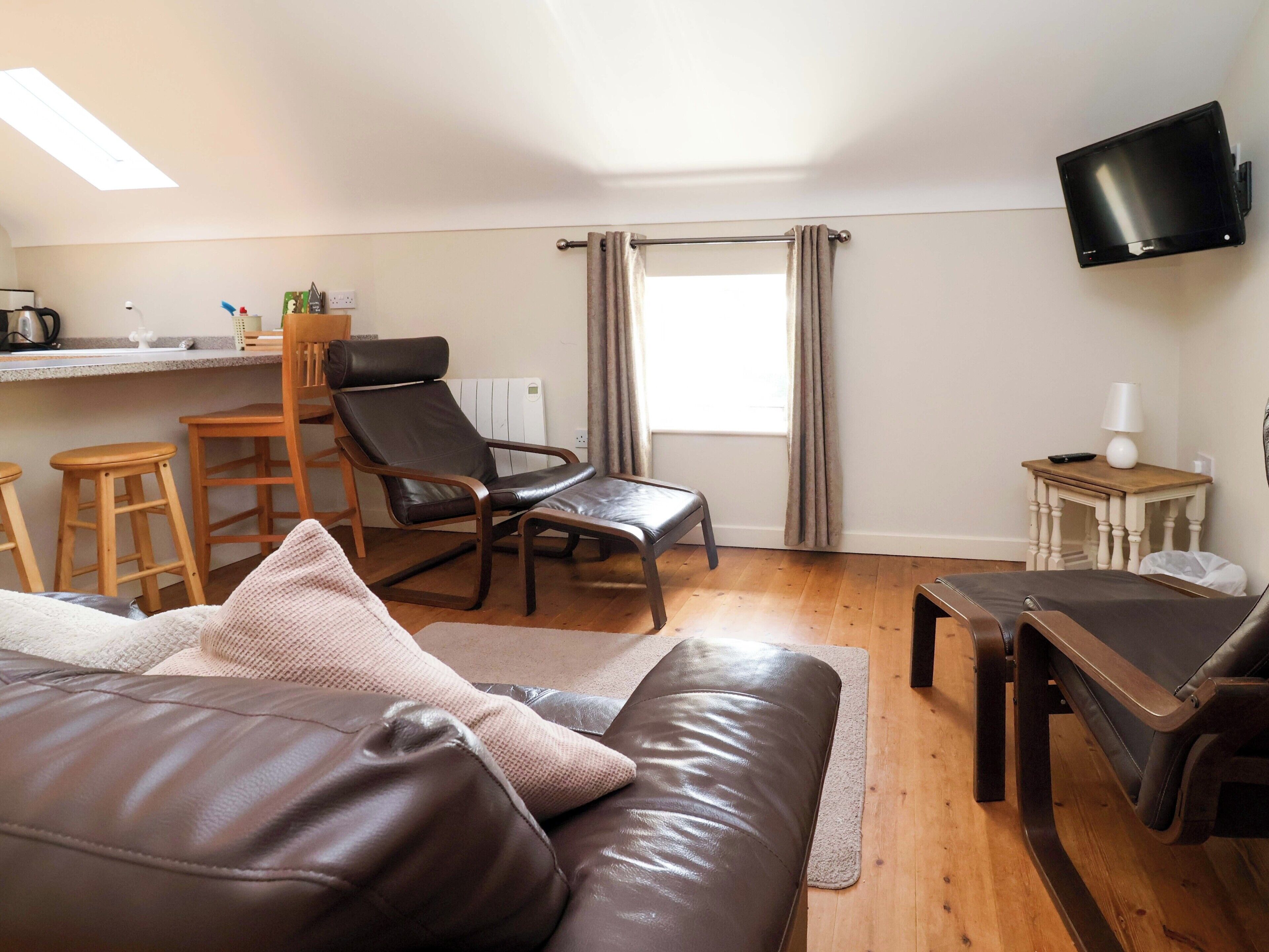 En kompakt opholdsstue med sofa, stol, lille bord, tv ophængt på væggen og et vindue med gardiner.