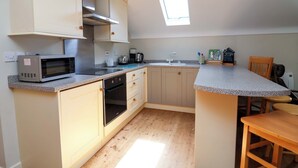 A compact kitchen with a microwave, oven, and sink. There is a small window providing natural light and a wooden dining table. - Pen Y Grisiau (Mold)