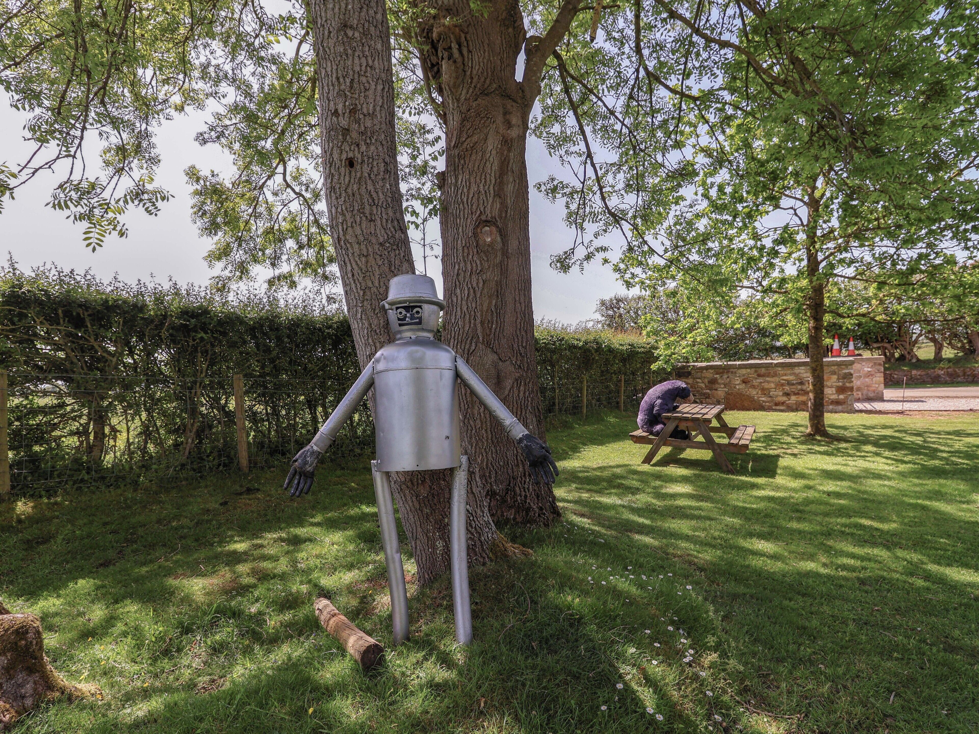 Escultura de una figura humanoide en un parque, con árboles y una mesa de picnic.