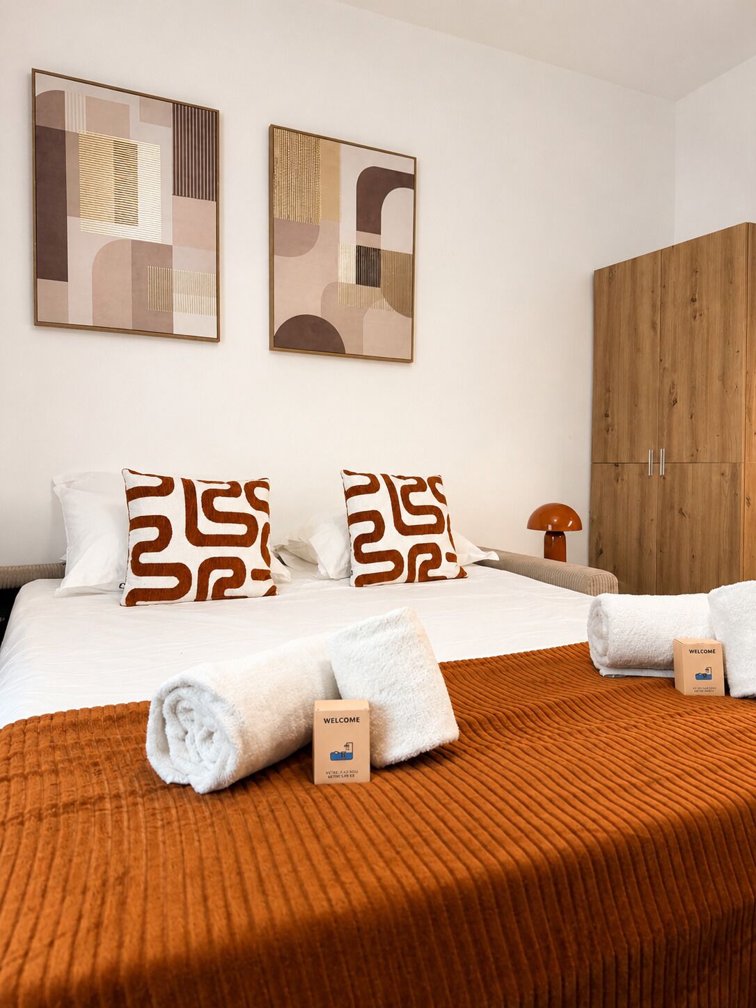 A hotel room with a bed, two pillows, a wooden wardrobe, a welcome box, and abstract wall art.