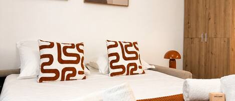A hotel room with a bed, two pillows, a wooden wardrobe, a welcome box, and abstract wall art.