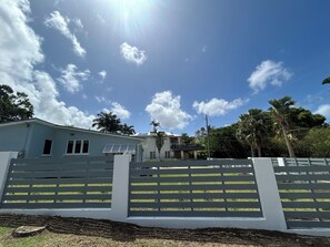 A residential property with a white fence, a house with a flat roof, and palm trees in the background. - 2 bed Villa in Hastings, Walking distance to Amenities - NO CAR NEEDED (Bridgetown)