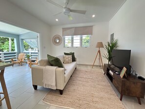 A living room with a sofa, a coffee table, a TV stand, and a floor lamp. There is a large window with a view of greenery outside. - 2 bed Villa in Hastings, Walking distance to Amenities - NO CAR NEEDED (Bridgetown)