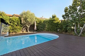 Una piscina con cubierta de madera, rodeada de vegetación y un edificio con techo de tejas.