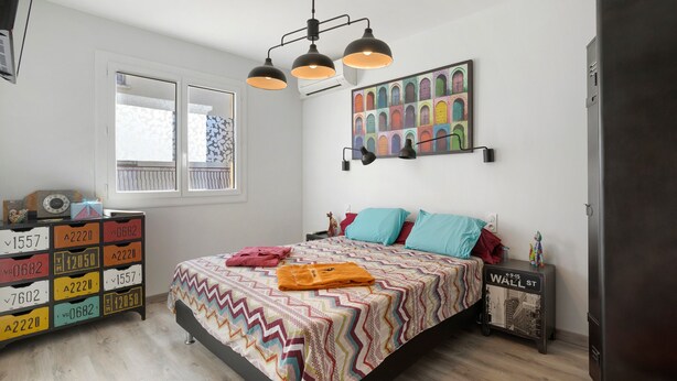 Un dormitorio con una cama, un aparador, una lámpara de pared, un cuadro de colores y una ventana.