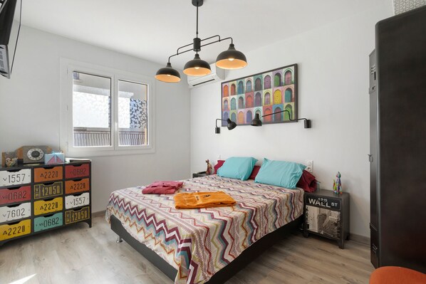 Un dormitorio con una cama, un aparador, una lámpara de pared, un cuadro de colores y una ventana.