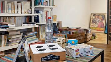 Una habitación con una estantería, una mesa con varios objetos, incluyendo una botella de agua, una caja con la etiqueta 'MAISON DE CAMPAGNE' y un mantel a rayas de colores.