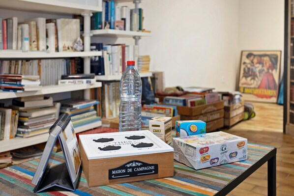 Una habitación con una estantería, una mesa con varios objetos, incluyendo una botella de agua, una caja con la etiqueta 'MAISON DE CAMPAGNE' y un mantel a rayas de colores.