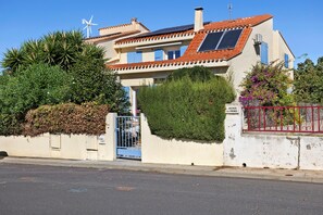 Una casa de dos pisos con una turbina eólica, paneles solares y un acceso con portón cerrado.