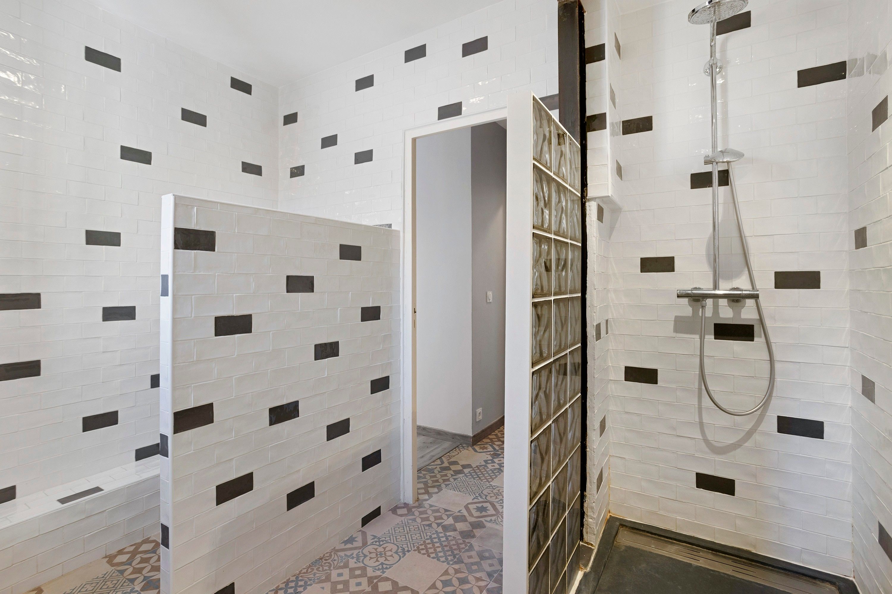 Une salle de bain avec des murs recouverts de carreaux de céramique blancs rectangulaires, une douche équipée d’un pommeau de douche amovible et un sol à motifs.