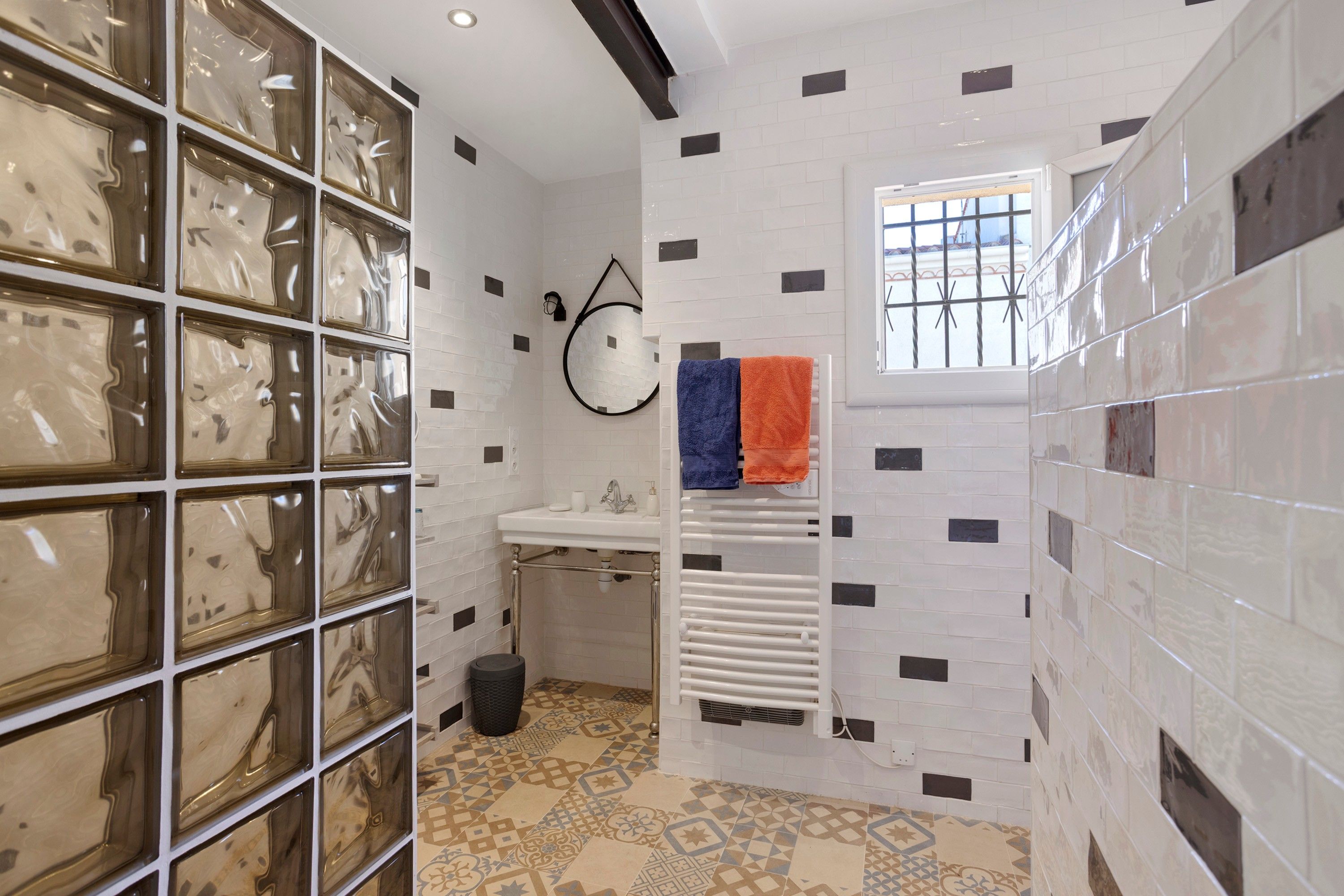 A bathroom with a white sink, a mirror, a towel rack with two towels, patterned floor tiles, and a window with bars.