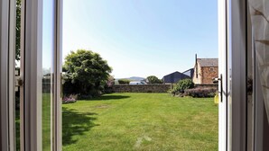 A view through a glass door of a grassy yard with a stone wall, a tree, and a building in the background. - Ty Haf (Mold)