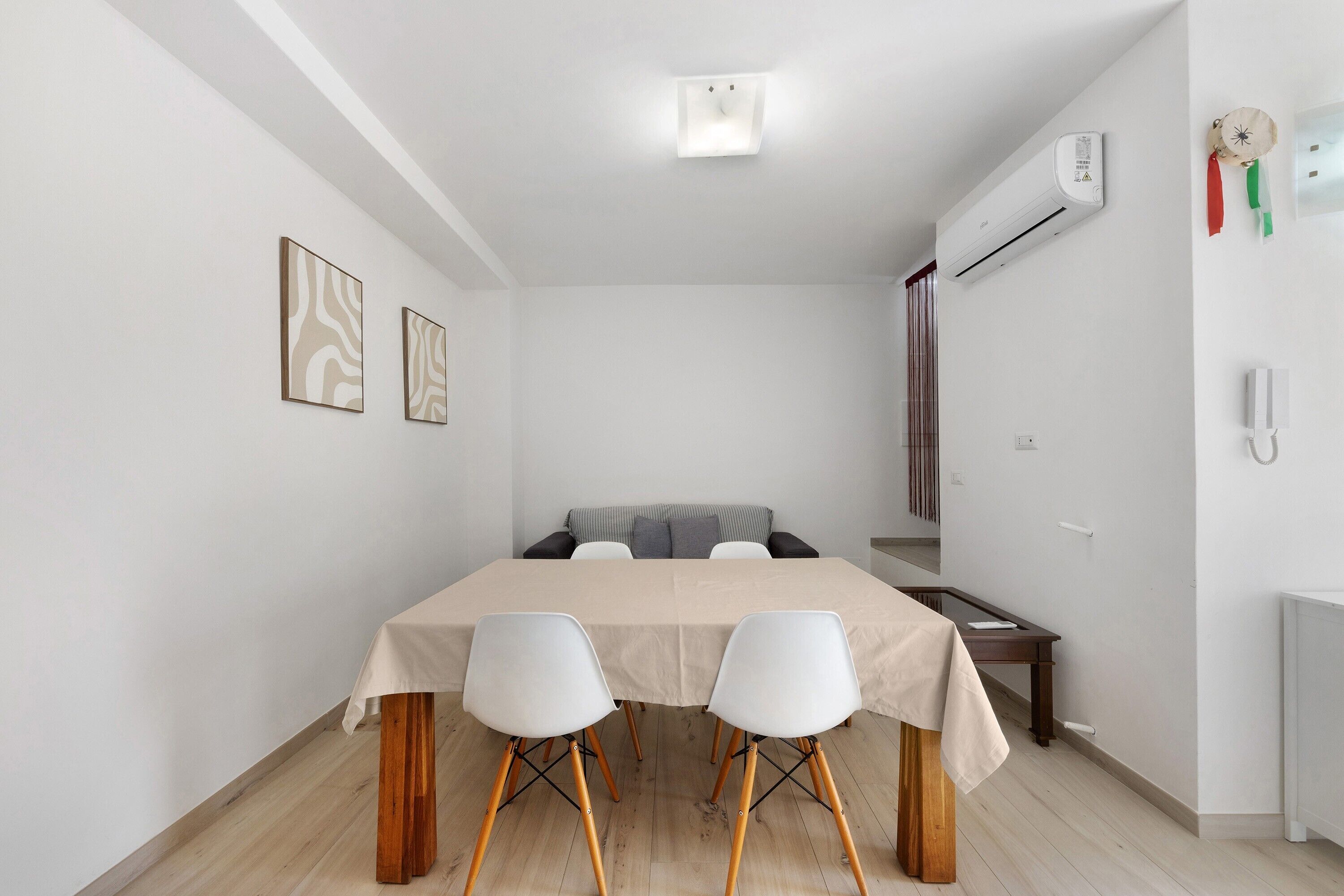A small, modern room with a bed, a table with two chairs, a bedside table, a wall-mounted air conditioner, and a wall-mounted telephone.