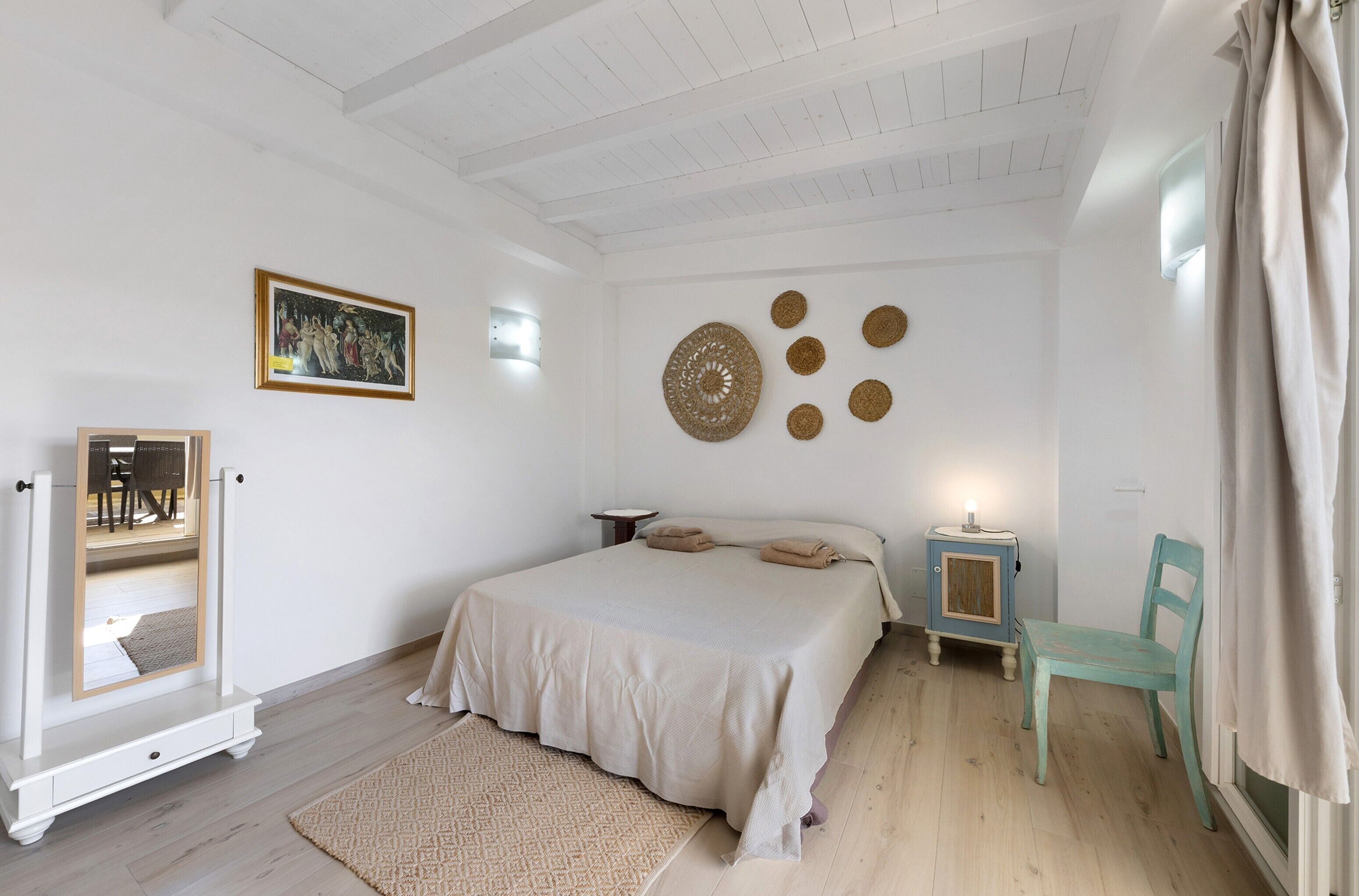 Un dormitorio con cama, una mesita de noche con lámpara, una silla, un espejo y una obra de arte en la pared.