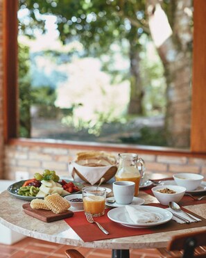 A breakfast setting with a variety of food items, including fruits, bread, and beverages, arranged on a table with a view of greenery outside. - Pousada do Cedro (Santo Antônio do Pinhal)