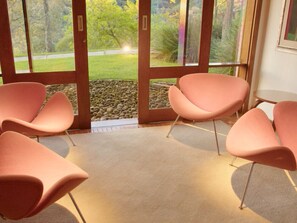 A room with four pink chairs arranged in a semi-circle, a carpeted floor, and a view through glass doors to an outdoor garden area with rocks and greenery. - Pousada do Cedro (Santo Antônio do Pinhal)