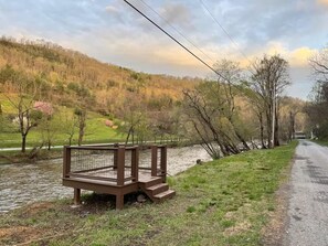 A wooden deck with a metal railing overlooking a river, surrounded by trees and a grassy area.