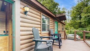A log cabin with a deck featuring two chairs and a table, a green door, and a car parked in the background. - Riverfront Wilderness Cabin (Cullowhee)