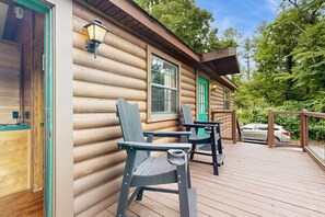 A log cabin with a deck featuring two chairs and a table, a green door, and a car parked in the background. - Riverfront Wilderness Cabin (Cullowhee)