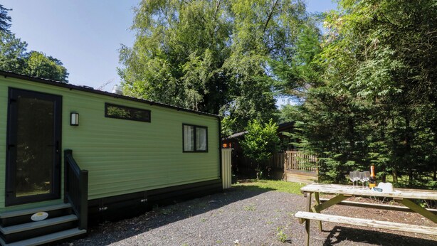 A green mobile home with a porch and steps, surrounded by trees and a picnic table. - High Perch (Windermere)